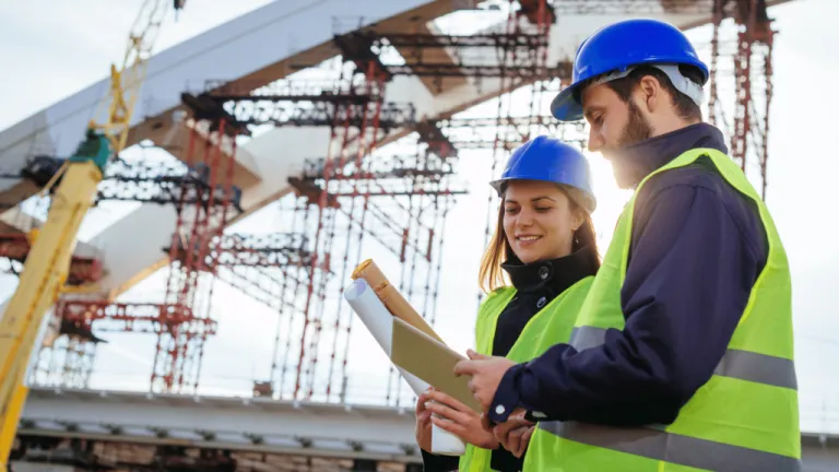 Engineers in hard hats reviewing plans in front of a bridge under construction